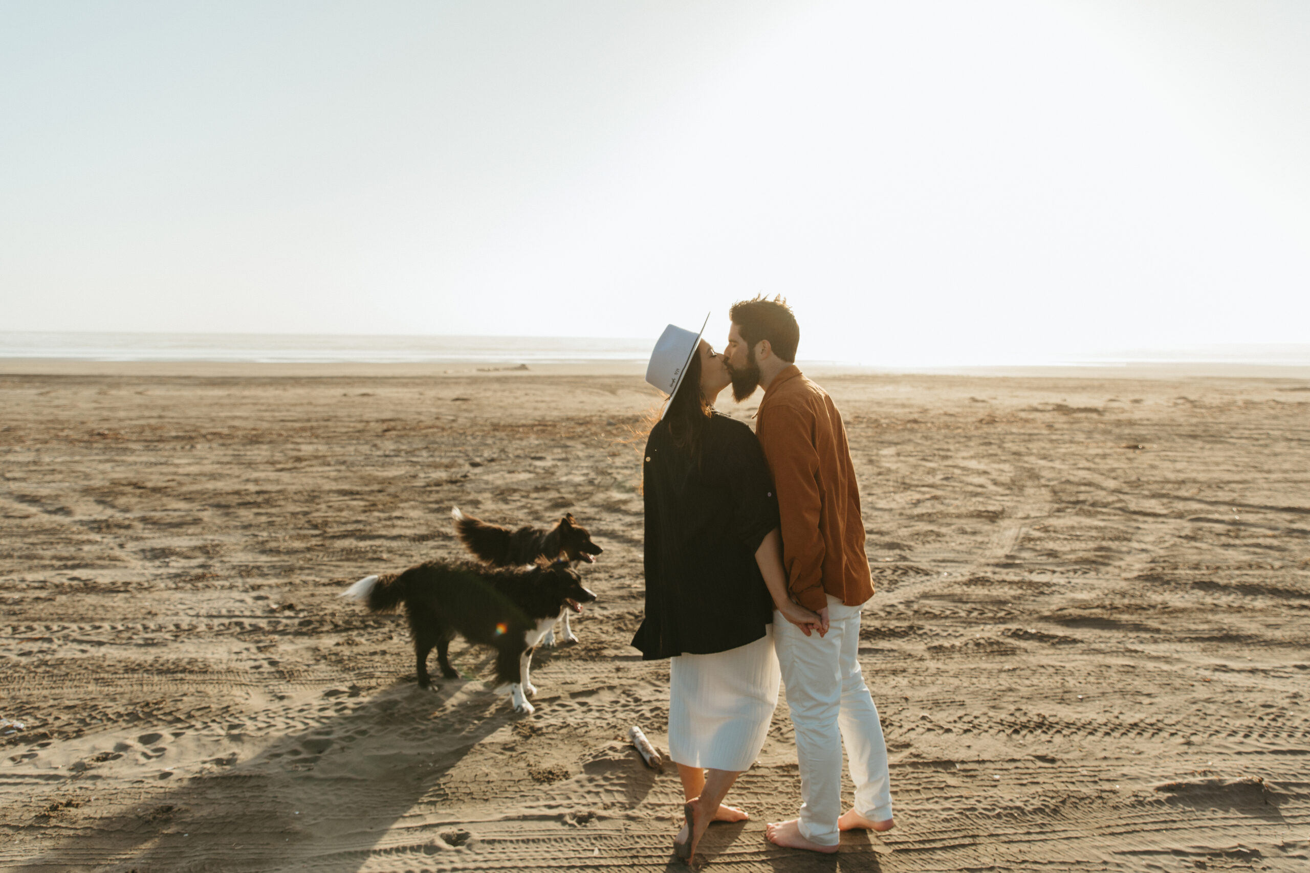 Couple at the Washington coast with their pups, kissing on the dunes in the sunset